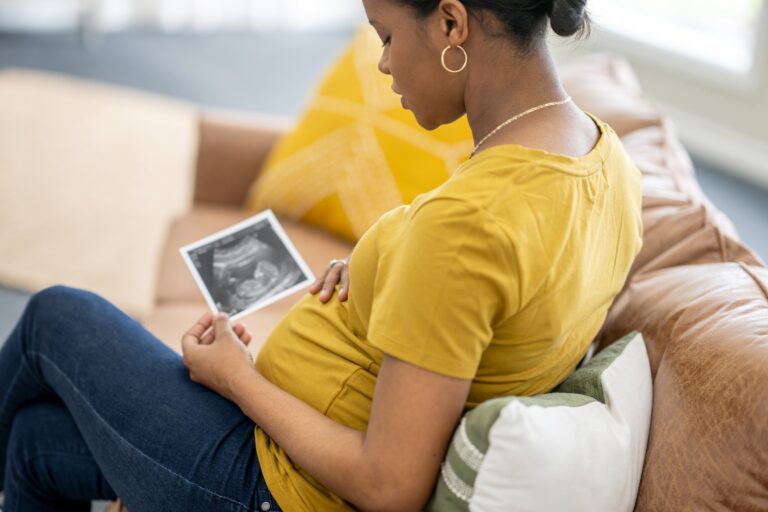 A young pregnant woman sits on a sofa in her home as she holds out an ultrasound picture