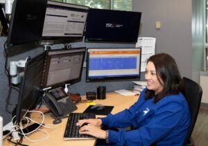 A registered nurse in blue scrubs sitting at a desk with several large monitors in front of her.